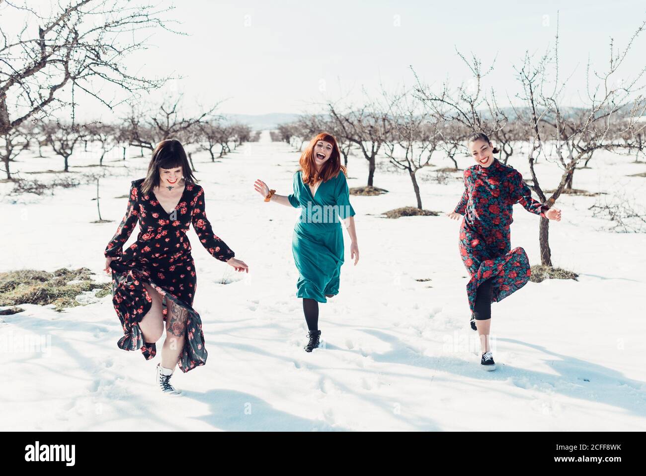 Fröhliche junge Freundinnen in bunten Kleidern mit Spaß und Laufen Auf Schnee im Winterfeld mit Baumbepflanzung Stockfoto