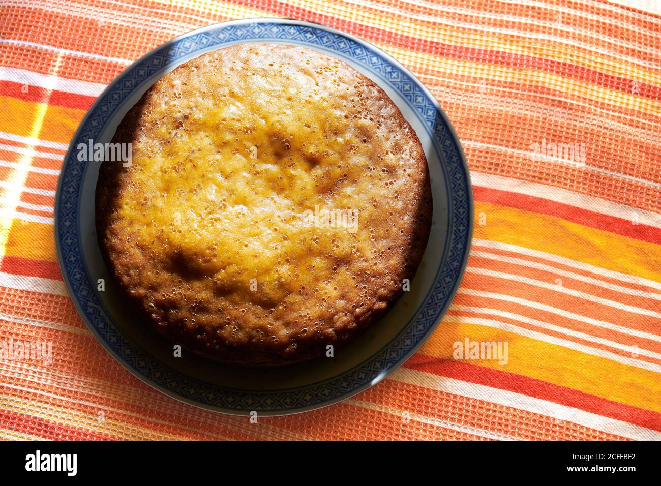 Frisch hausgemachter Bananenkuchen mit gelber Kruste auf einem Teller auf einem bunten Tisch präsentiert. Die Sonne scheint auf dem Kuchen. Stockfoto