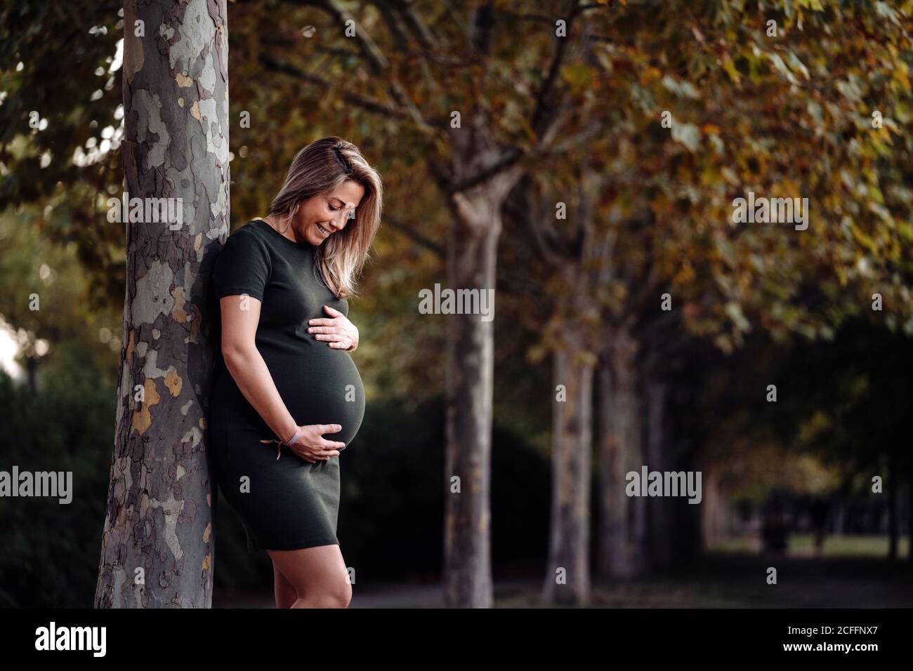 Glückliche Schwangere in lässigem Kleid streichelte Bauch, während sie an sonnigen Sommertagen auf einem grünen Baum mit blauem Himmel stand Stockfoto