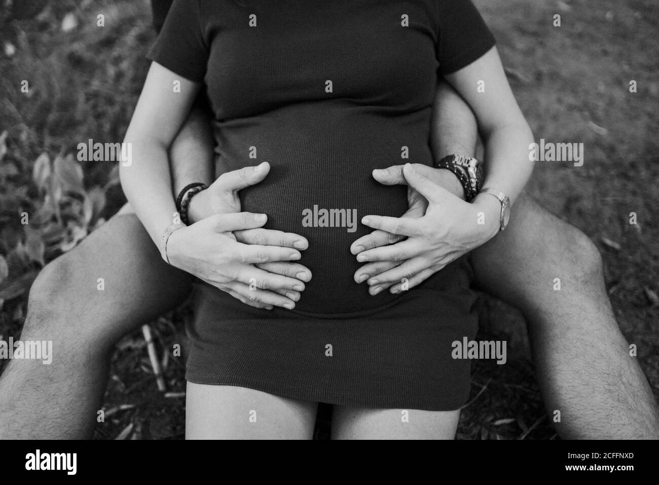 Crop Mann umarmt schwangere Frau und beide halten Hände auf Bauch beim Sitzen zusammen auf Gras im Park Stockfoto