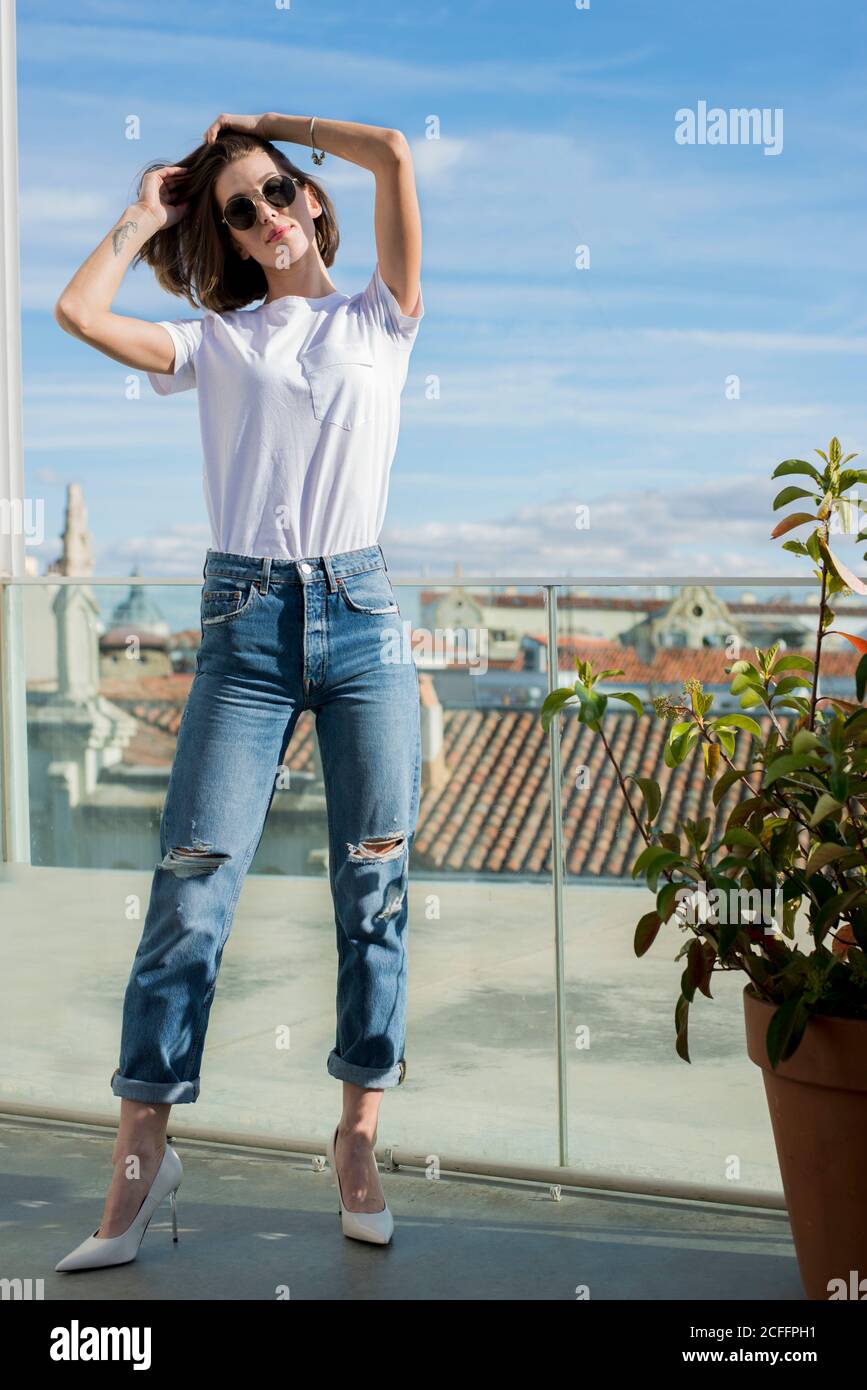 Slim weibliche Modell in Jeans und High Heel Schuhe stehen Mit geneigtem Kopf und berühren Haare in der Nähe transparente Trennwand in Tageslicht Stockfoto