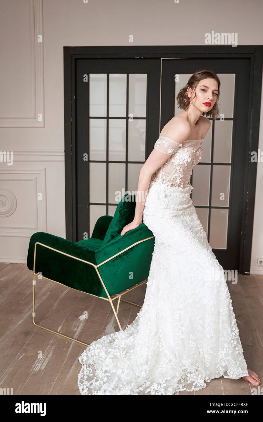 Barfuß schlanke Braut in eleganten weißen Kleid Blick auf die Kamera Während er auf der Rückenlehne des grünen Sessels gegen schwarze Türen sitzt In geräumigen Wohnung Stockfoto