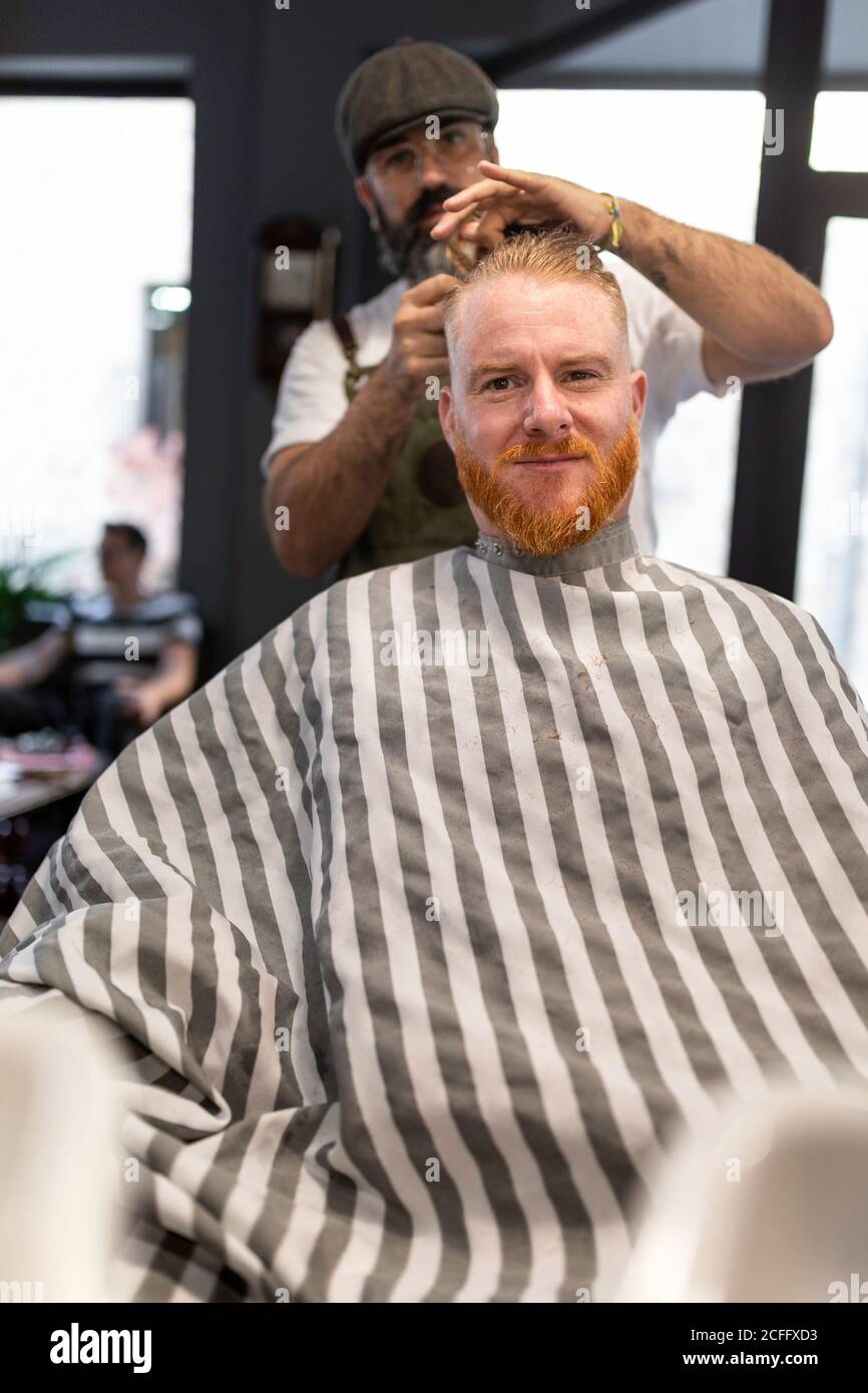 Moderner Friseur Coiffeur schneidet einem erwachsenen Mann Rothaarhaare in Friseurstuhl Stockfoto