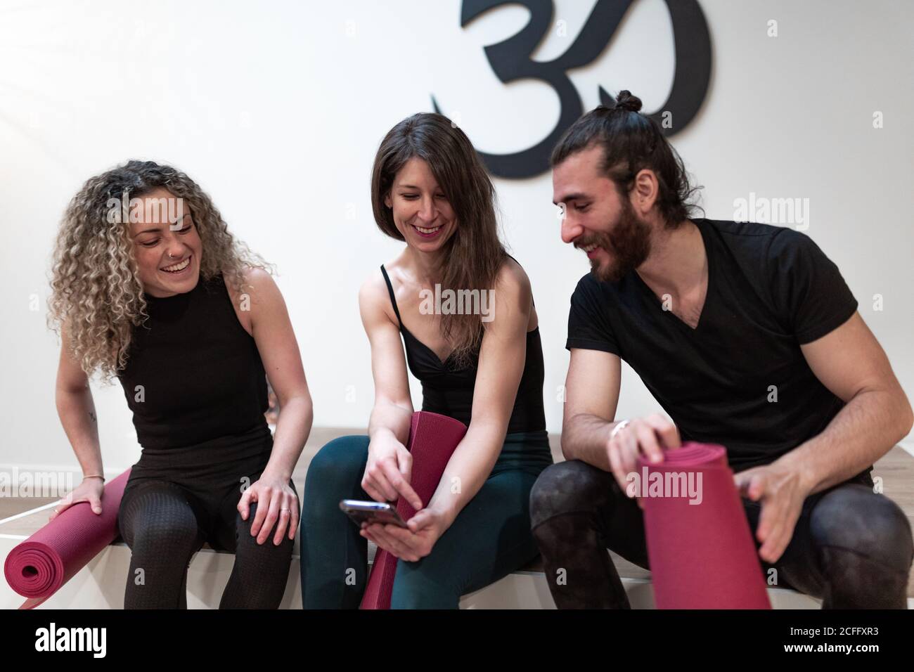 Glückliche junge Frau lächelt und zeigt den begeisterten Freunden mit Matten vor dem Yoga-Training im Studio Daten auf dem Smartphone Stockfoto