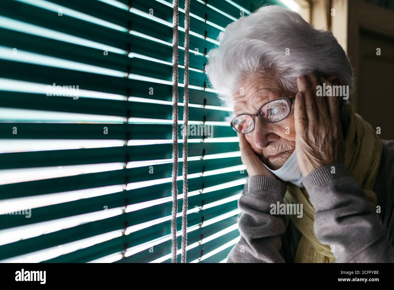 Unglückliche einsame ältere Frau mit Schutzmaske, die durch zugemacht schaute Fenster während des Aufenthalts zu Hause während des Coronavirus-Ausbruchs Stockfoto