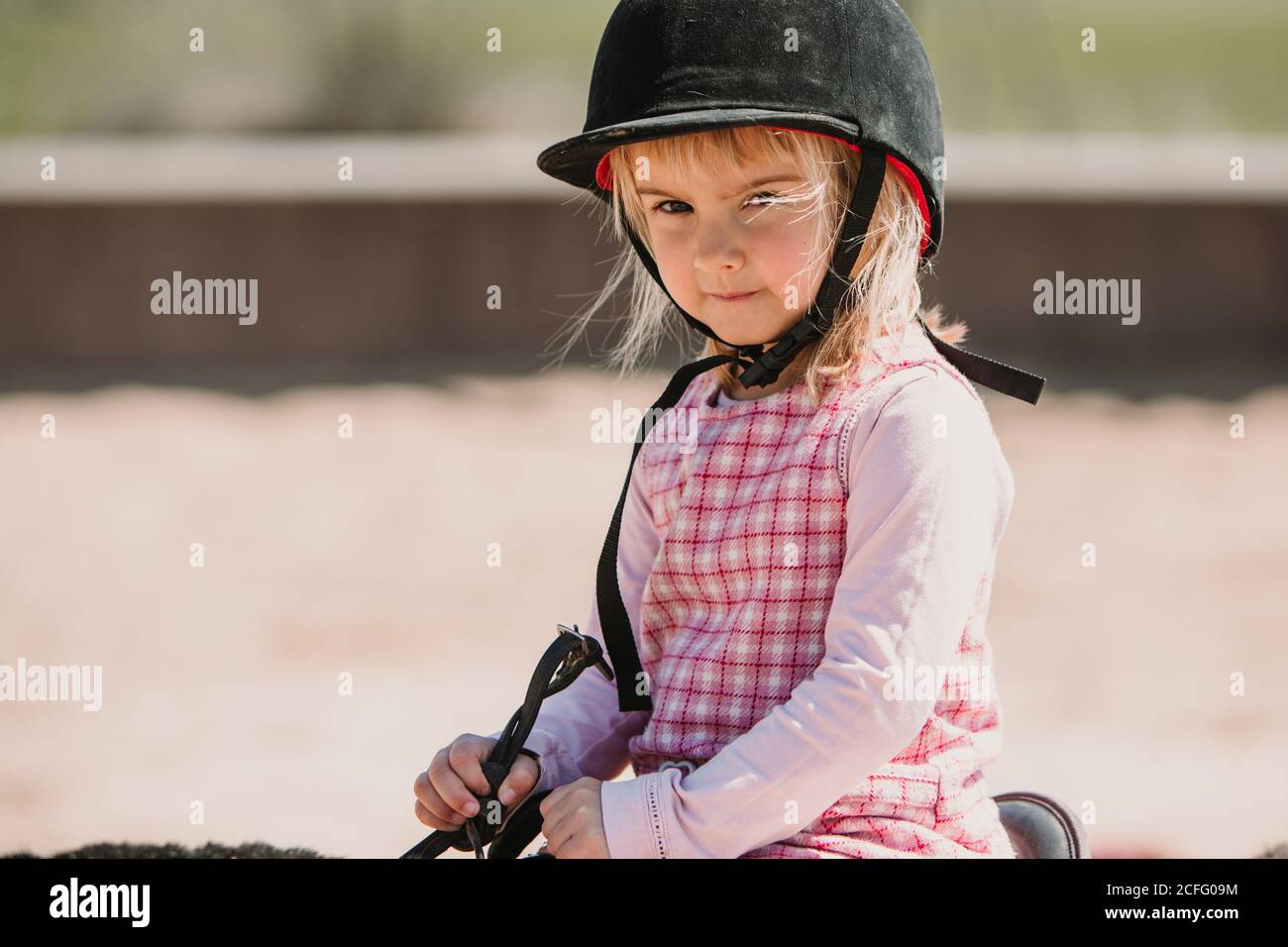 Kleines Mädchen in Kleid und Jockey Heu sitzt auf Pferd Während des Lernens auf der Rennstrecke zu fahren Stockfoto
