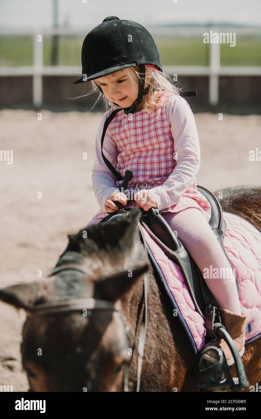 Kleines Mädchen in Kleid und Jockey Heu sitzt auf Pferd Während des Lernens auf der Rennstrecke zu fahren Stockfoto