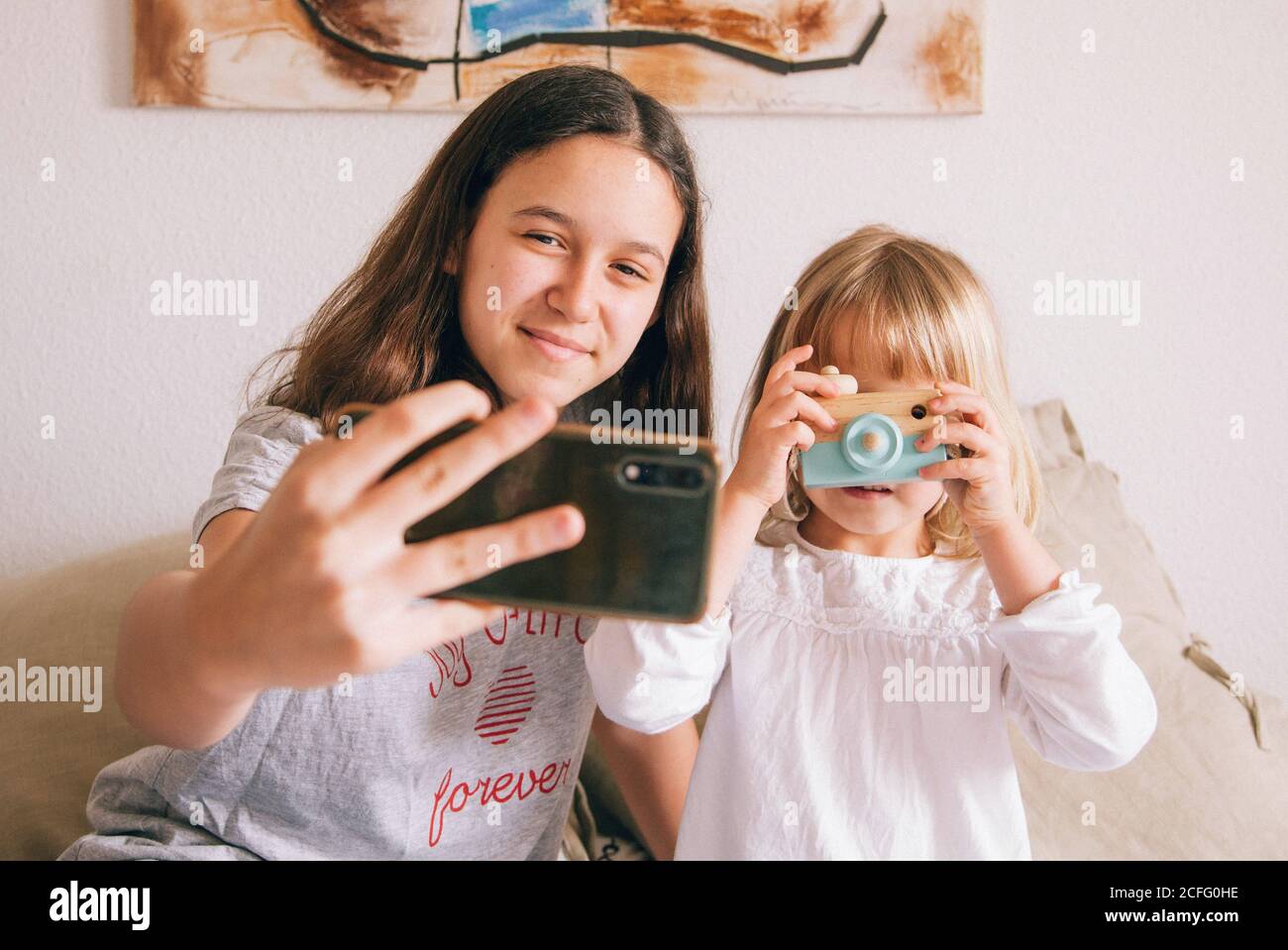 Charmante Teenager-Mädchen mit und kleine Schwester sitzen auf komfortabel Bett und Fotos auf Smartphone und Spielzeugkamera Stockfoto