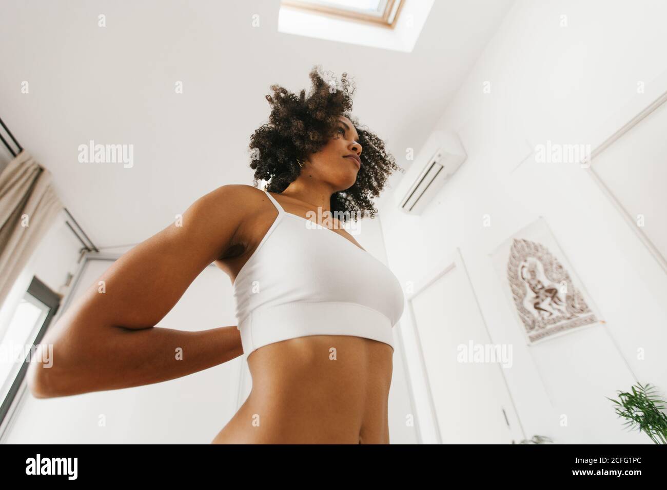 Von unten afroamerikanische attraktive junge Frau, die Yoga-Haltung mit gefalteten Händen hinter dem Rücken in hellen Raum Stockfoto