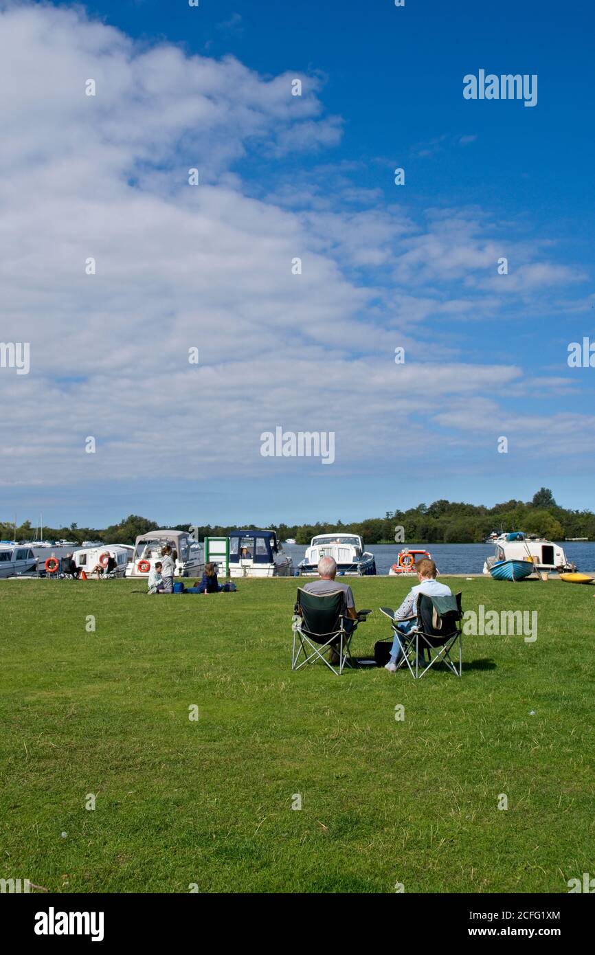 Bootsanlegestellen auf den Broads. Paar sitzen in Camper Stühle auf Gras Patch beobachten Bootfahren Aktivität in Pub Liegeplätze. Leichte Wolke in blauem Himmel. Stockfoto