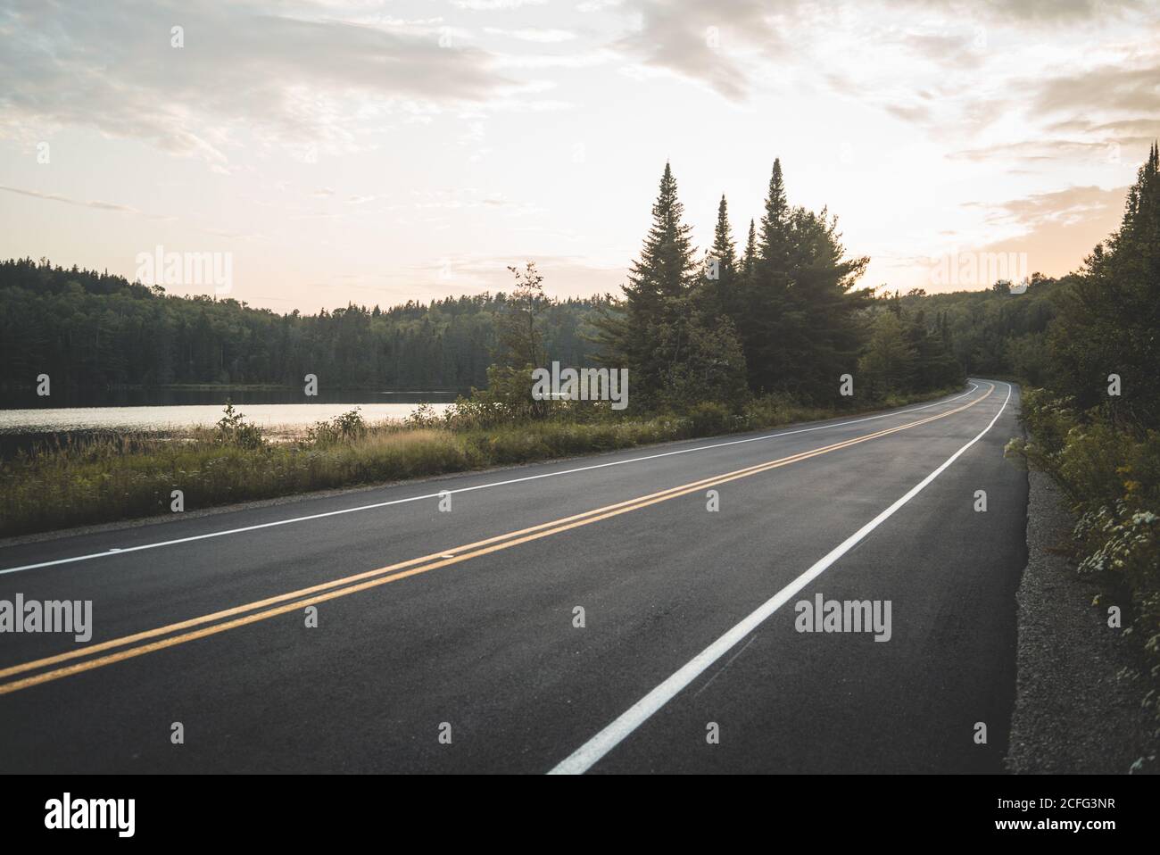 Asphaltstraße in der Nähe von ruhigen See und grünen Wald gegen bewölkten Sonnenuntergang Himmel in La Mauricie Nationalpark in Quebec, Kanada Stockfoto