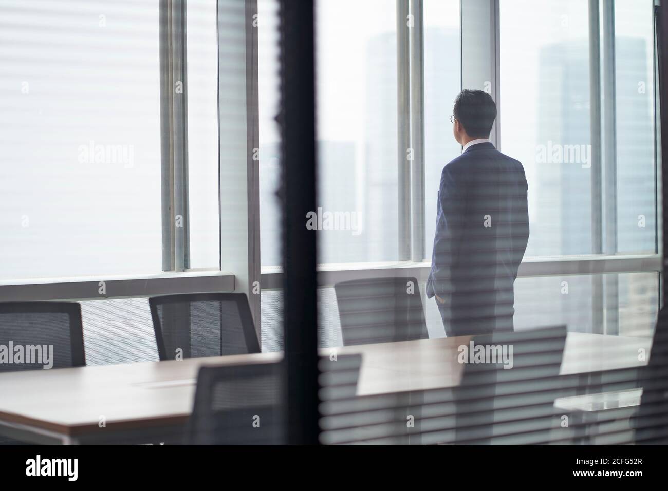 Rückansicht eines asiatischen Geschäftsmanns, der im Zimmer stand Vor dem Bürofenster mit Blick auf die Tür und überlegt Tasche Stockfoto