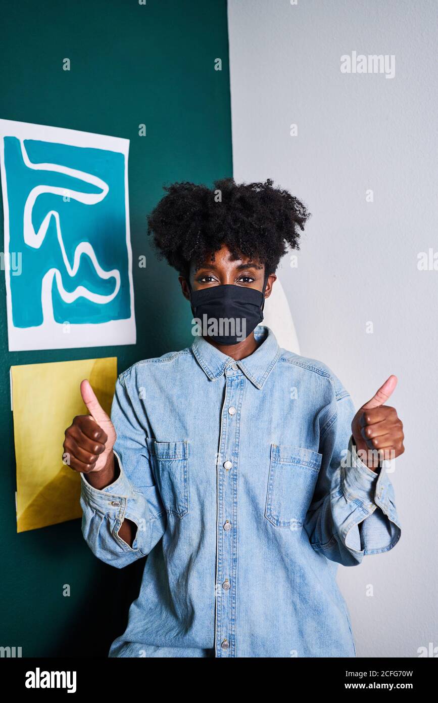 Junge afroamerikanische Frau in legerem Denim-Hemd und Schwarz Schutzmaske, die auf die Kamera schaut und die Daumen nach oben zeigt Während des Aufenthalts zu Hause während des Coronavirus-Ausbruchs Stockfoto