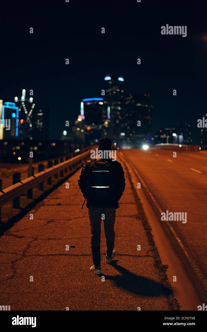 Rückansicht des anonymen Mannes in legerer schwarzer Lederjacke Mit Rucksack zu Fuß in der Nacht Stadt auf verschwommenem Hintergrund Stockfoto