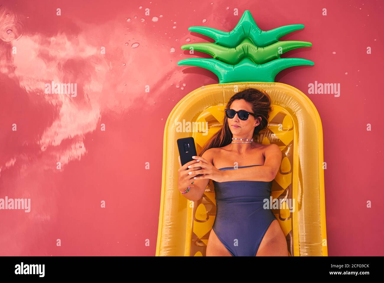 Von oben Frau in Sonnenbrille und Badeanzug auf Ananas Luftmatratze liegend Selfie auf rosa Wasser von Salzsee Stockfoto