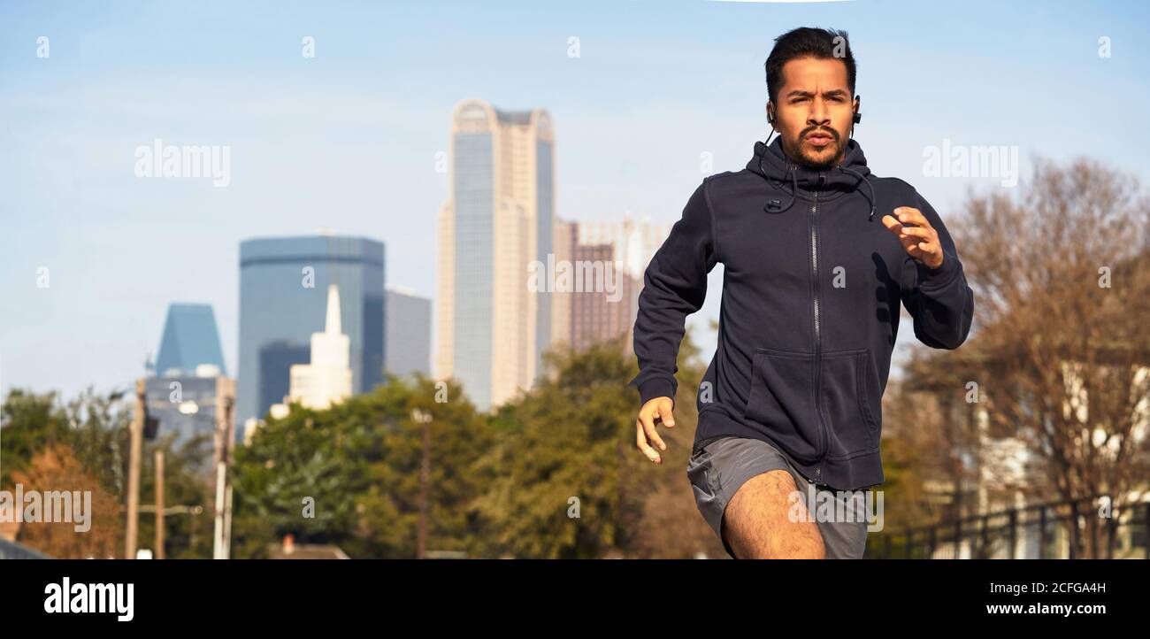 Konzentrierter hispanischer männlicher Athlet in Sportbekleidung, der mit wegschaut Kopfhörer laufen auf leerer Straße in der Innenstadt von Dallas Stockfoto
