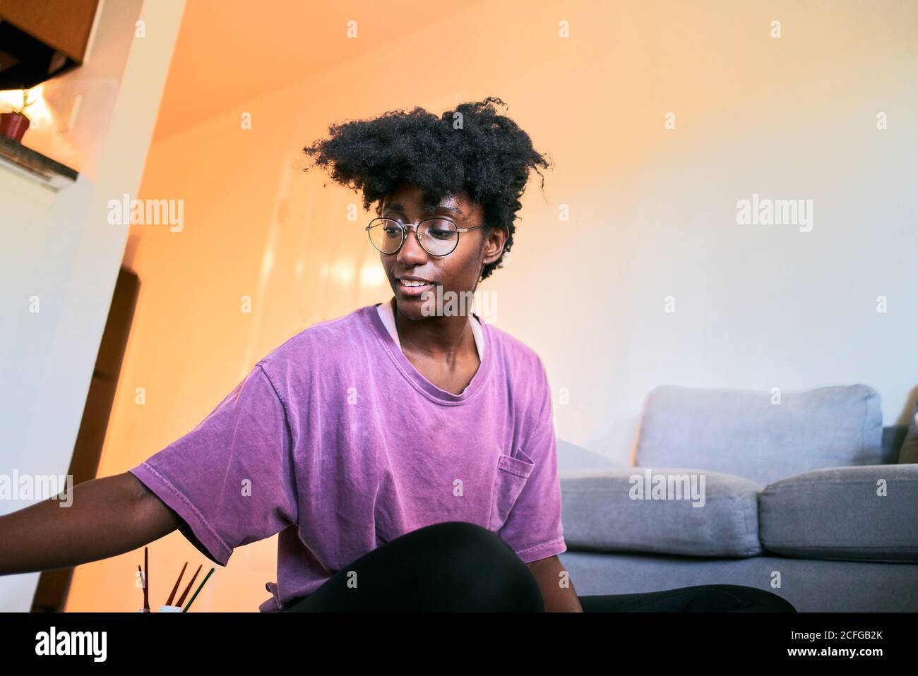 Positive junge afroamerikanische Frau mit Afro-Haar und Brillen Moderne Gläser in legerer Kleidung auf dem Boden sitzen und malen Mit Bürsten, während Sie Zeit zu Hause während Ihres Aufenthalts verbringen Home Kampagne für Coronavirus Prävention Stockfoto