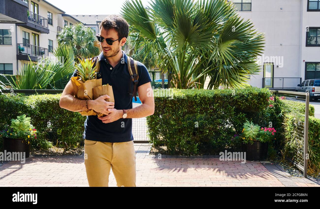 Fröhlicher Mann in Sonnenbrille im trendigen Outfit auf der Straße stehend Mit Einkaufstaschen, die wegschauen Stockfoto