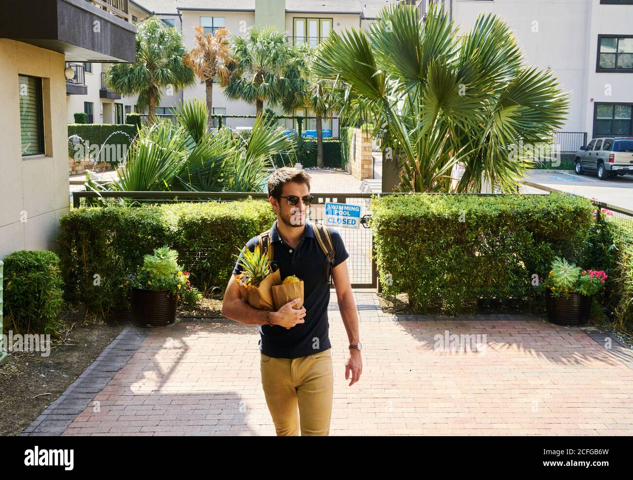Fröhlicher Mann in Sonnenbrille im trendigen Outfit auf der Straße stehend Mit Einkaufstaschen, die wegschauen Stockfoto