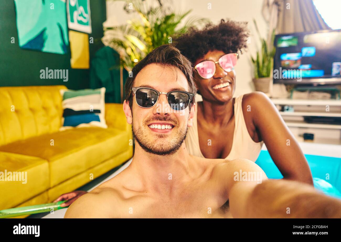 Fröhliche multirassische Paar trägt Sonnenbrille sitzt in aufblasbaren Pool und Selfie beim Genießen des Wochenendes Stockfoto