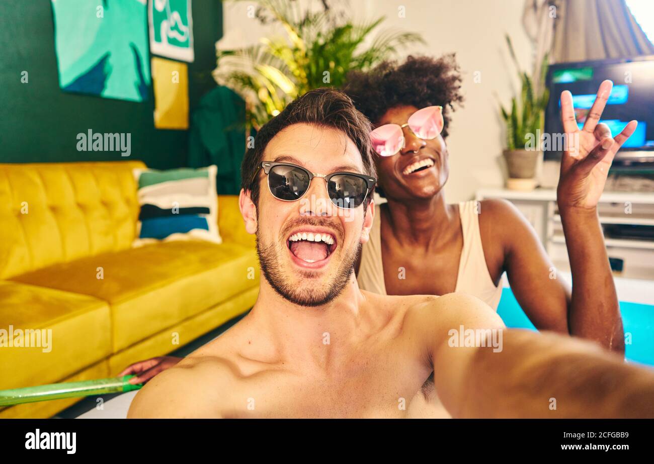 Fröhliche multirassische Paar trägt Sonnenbrille sitzt in aufblasbaren Pool und Selfie beim Genießen des Wochenendes Stockfoto