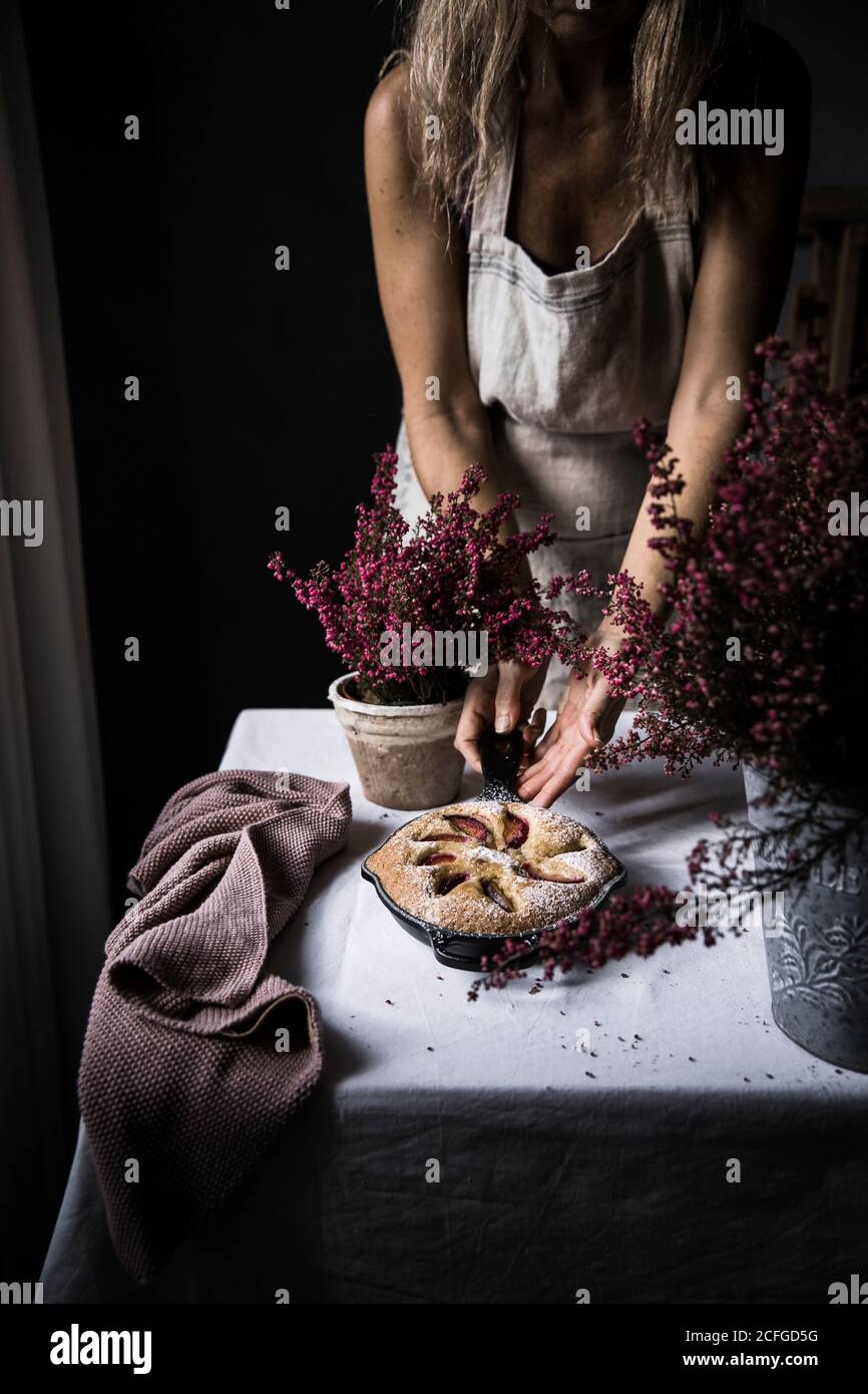 Crop Frau kochen in Schürze schneiden Pflaumenkuchen auf Tisch mit Blumen Stockfoto