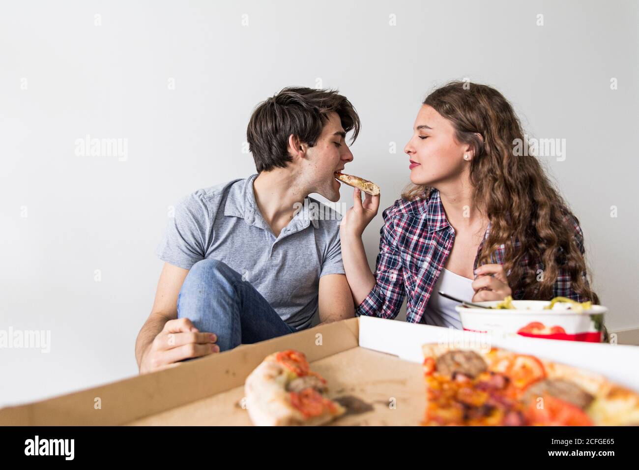 Niedliches junges Paar, das auf dem Boden sitzt und Pizza im Karton isst Kasten neben Leiter und lachend Stockfoto