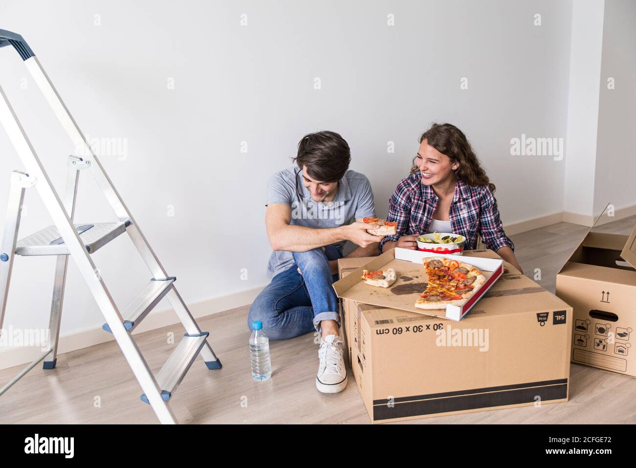 Niedliches junges Paar, das auf dem Boden sitzt und Pizza im Karton isst Kasten neben Leiter und lachend Stockfoto