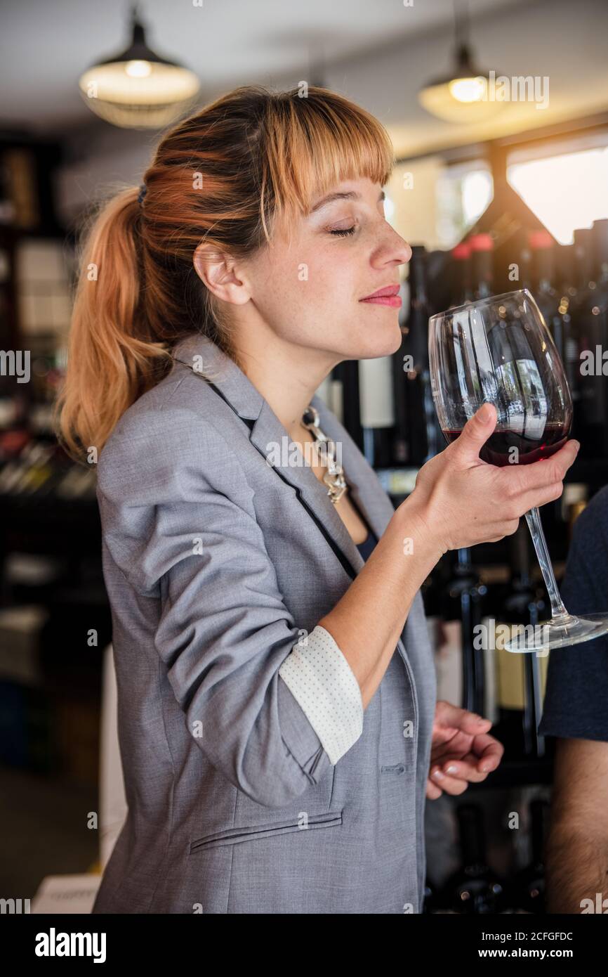 Seitenansicht einer eleganten Frau in einer Jacke, die Rotwein verkostet und ihn mit geschlossenen Augen im winehouse riecht. Stockfoto