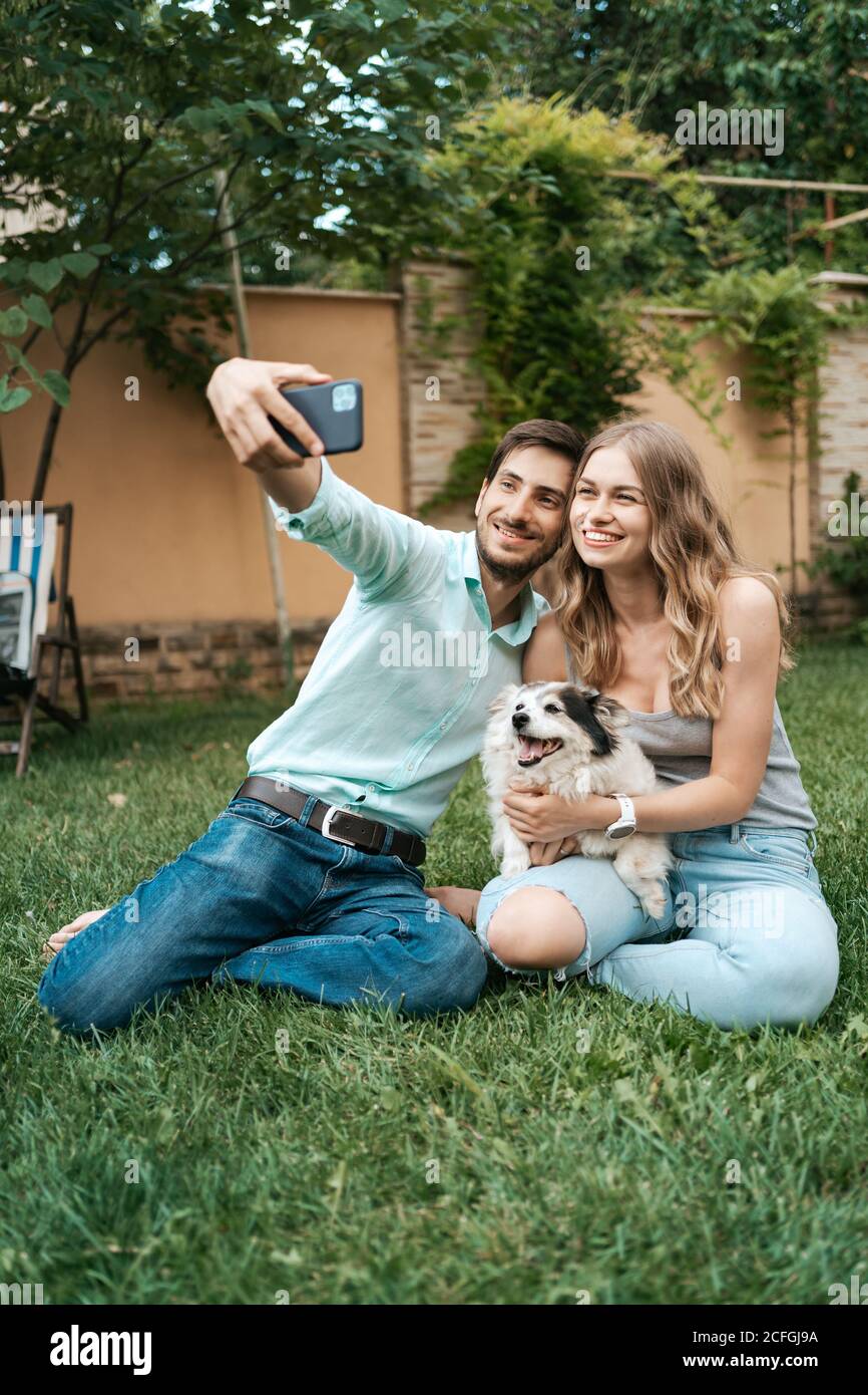 Schöne glückliche Paar machen Selfie mit ihren schönen Hund auf Der Hinterhof, während Sie auf dem Gras sitzen Stockfoto