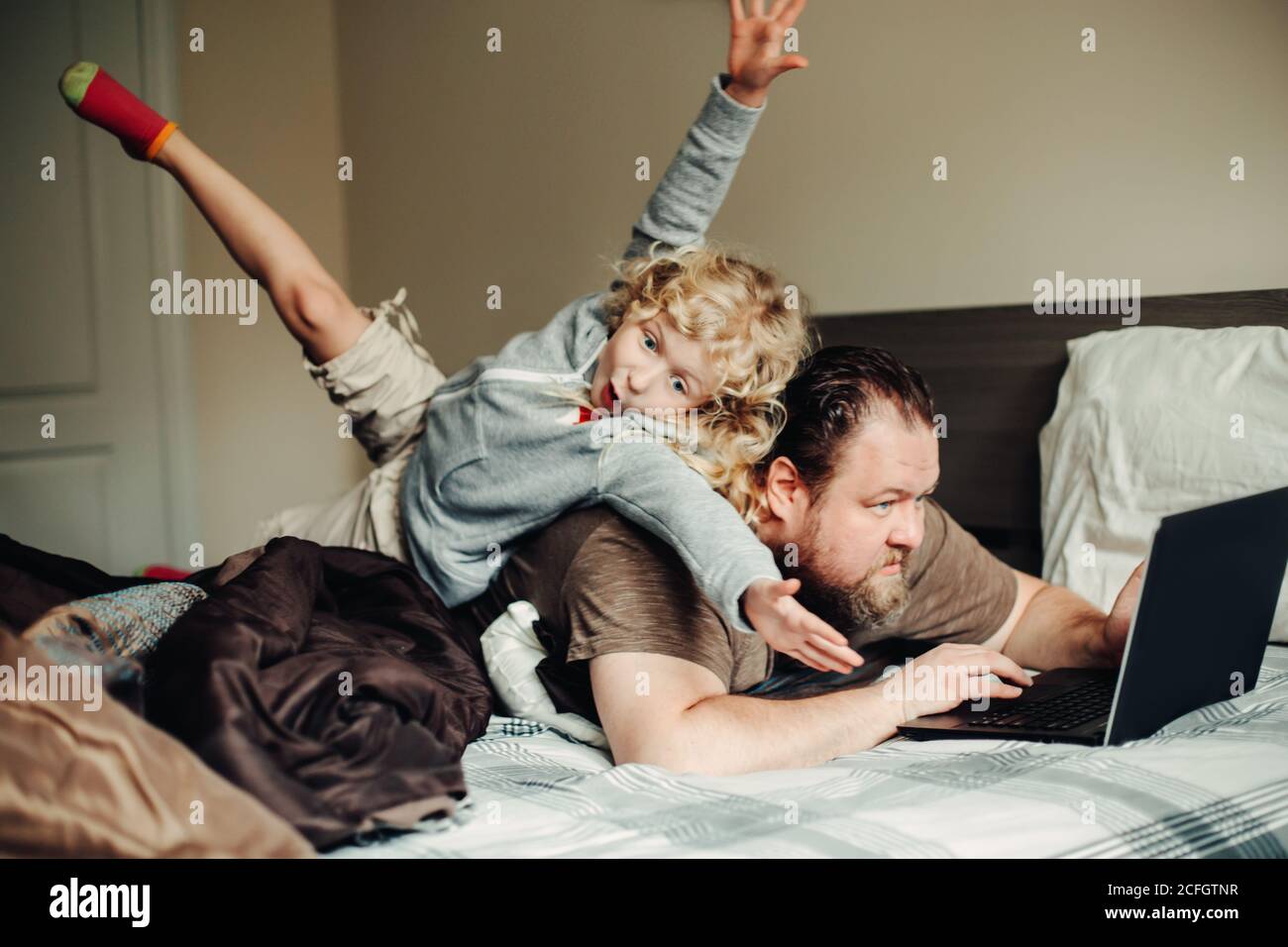Arbeiten Sie von zu Hause aus mit Kindern. Vater arbeitet am Laptop im Schlafzimmer mit Kind Tochter auf dem Rücken. Lustige ehrliche Familie Moment. Neuer Normalwert während Stockfoto