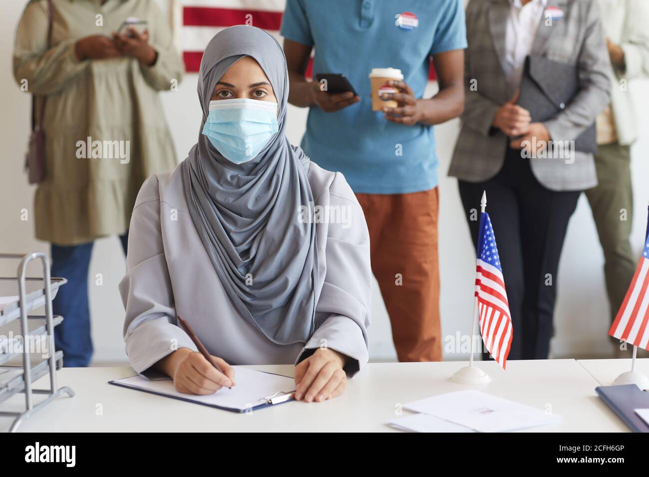 Multiethnische Gruppe von Menschen, die am Wahltag in einer Reihe stehen und Masken in der Wahlstation tragen, konzentrieren sich auf junge arabische Frau, die bei der Registrierung für die Wahl auf die Kamera schaut, Kopierraum Stockfoto