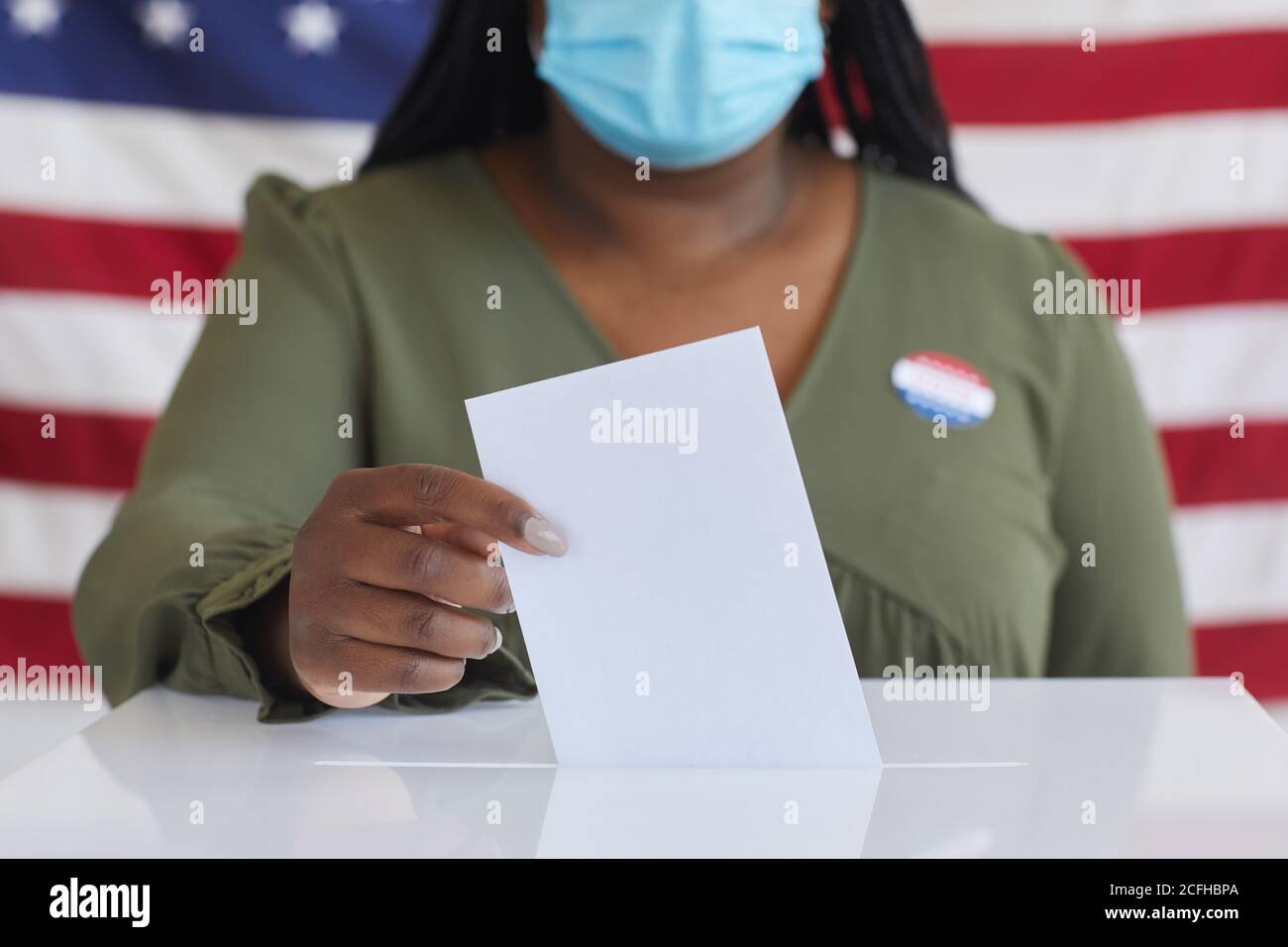 Nahaufnahme einer afroamerikanischen Frau, die eine Maske trägt, die ein Stimmzettel in die Wahlurne legt und die Kamera anschaut, während sie am Wahltag gegen die amerikanische Flagge steht, Platz kopieren Stockfoto