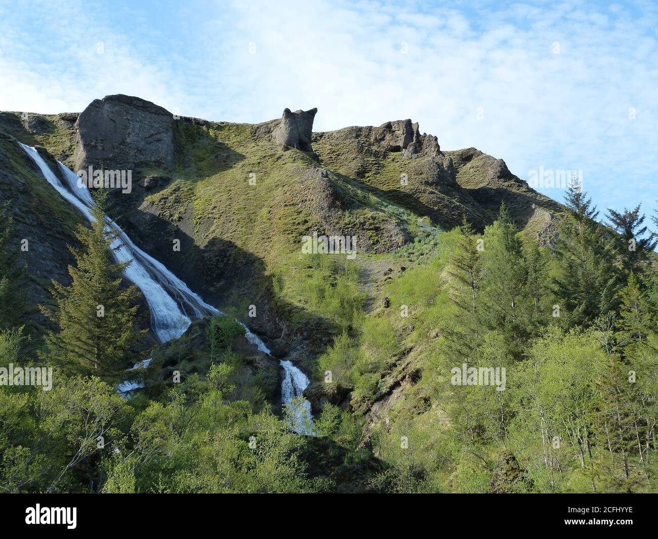 Schöne Sommer grüne Landschaft in Island. Nadelwald, Berge, Wasserfall. Wolkiger blauer Himmel. Erstaunlicher Wasserfall Systrafoss auf grünem Hügel. Stockfoto