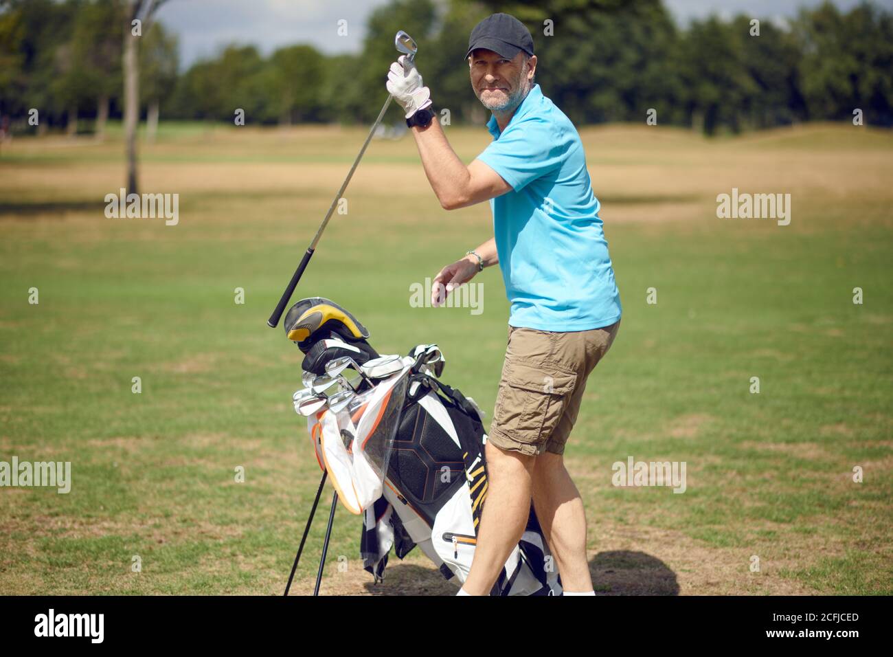 Mann mittleren Alters spielt eine Runde Golf auf einem Golf Kurs dreht sich, um die Kamera zu lächeln, während er auswählt Ein Club aus seiner Tasche für den nächsten Schuss Stockfoto