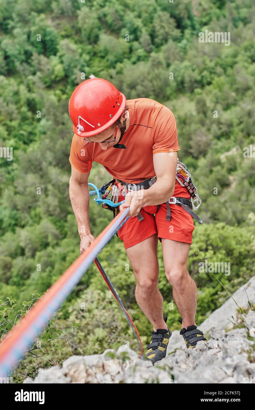 Von oben tapferer männlicher Alpinist in Schutzausrüstung Klettern Steile Klippe auf dem Hintergrund des Waldes Stockfoto