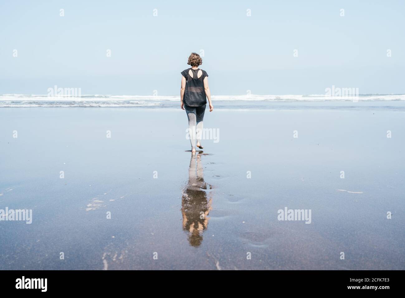 Ganzkörper-Rückansicht von nicht erkennbaren barfuß-Weibchen in Sportbekleidung Wandern auf nassem Sand in Richtung winkendes Meer Stockfoto