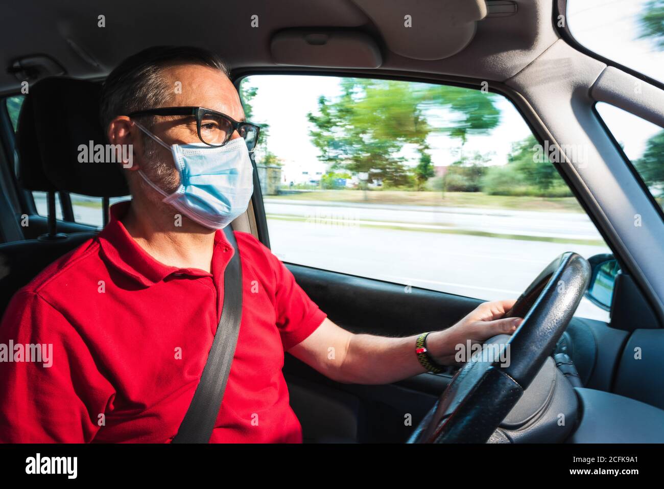 Älterer Mann im mittleren Alter in medizinischer Maske fährt modernes Auto Auf der Straße, während Sie sich nach vorne freuen Stockfoto
