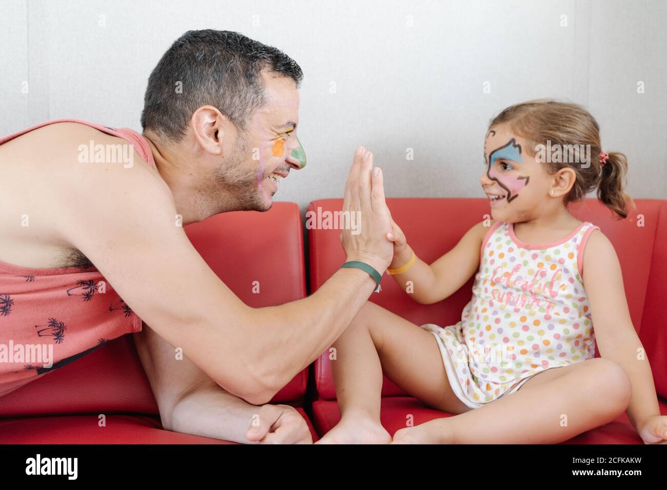 Seitenansicht von lächelnden Mann und kleines Mädchen mit lustig Bemalte Gesichter entspannen auf dem Sofa zu Hause, während hoch Fünf und einander anschauend Stockfoto