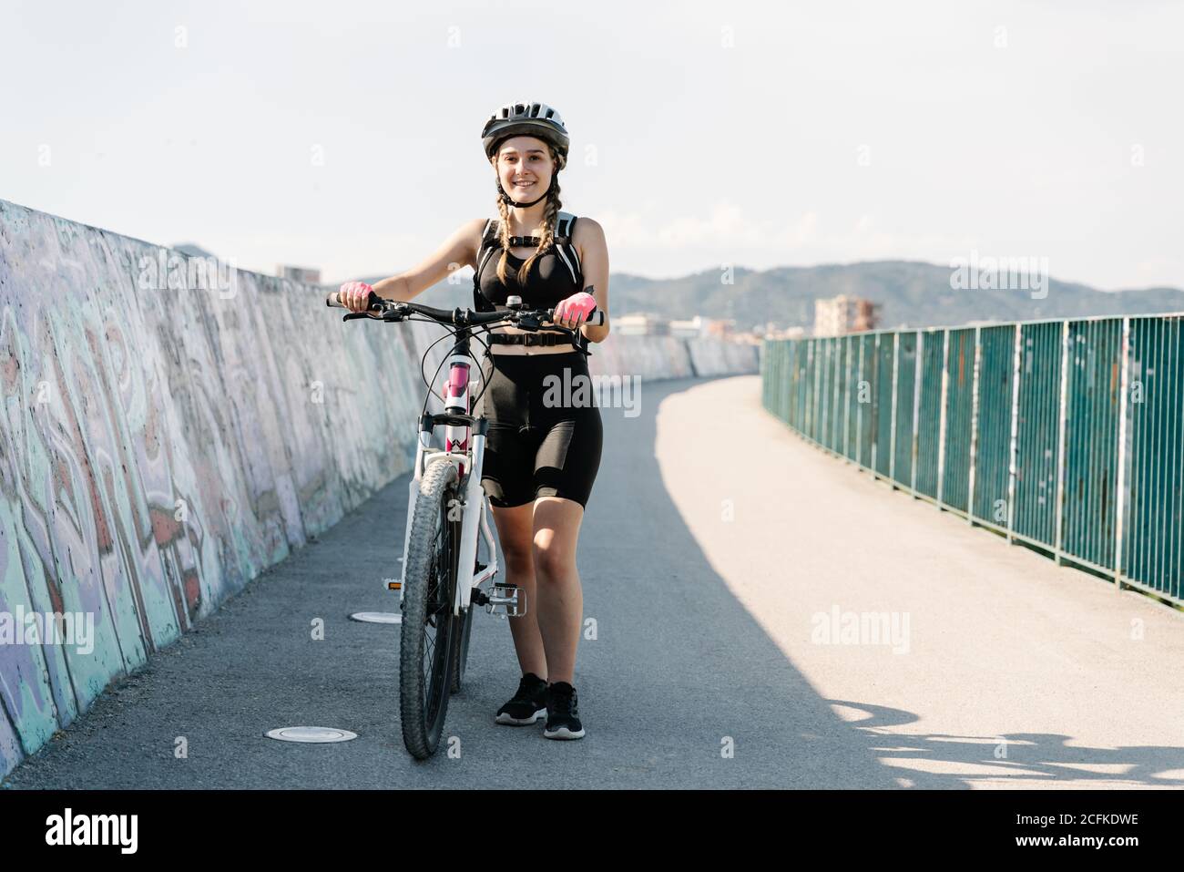Positive junge Radfahrerin in schwarzer Sportbekleidung und Schutzhelm Stehen mit dem Fahrrad auf asphaltierter Straße Stockfoto