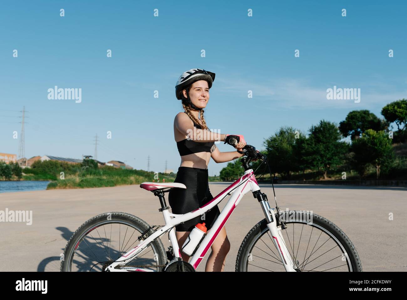 Seitenansicht einer positiven jungen Radfahrerin in schwarzer Sportbekleidung Und Schutzhelm stehen mit Fahrrad auf asphaltierter Straße und Selbstbewusst wegschauen Stockfoto
