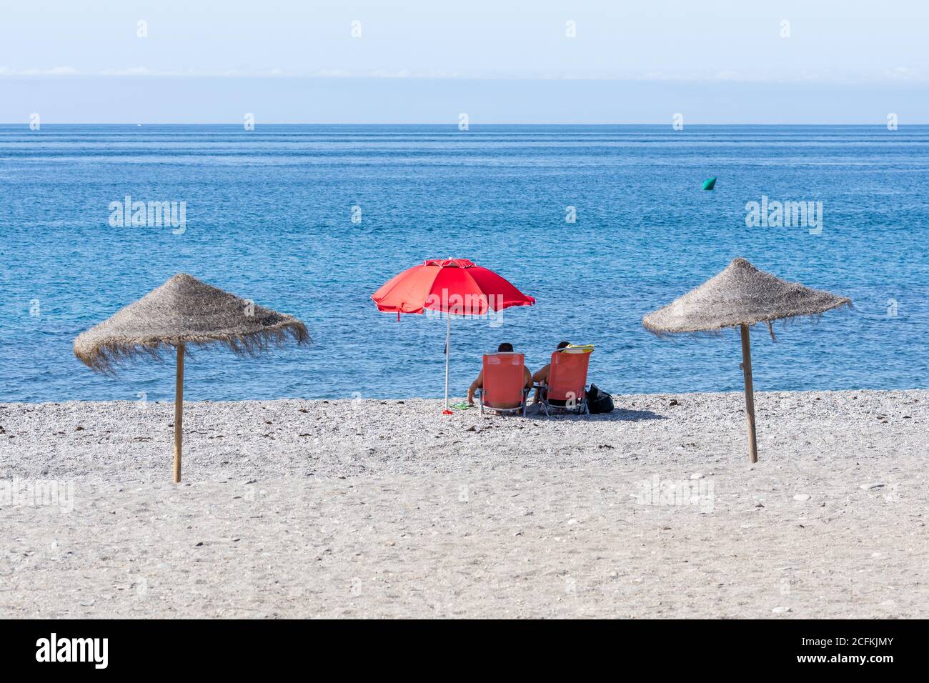 Andalusischer Strand ein sonniger Sommermorgen mit zwei Personen sitzen Unter einem roten Sonnenschirm mit Blick auf das Meer Stockfoto