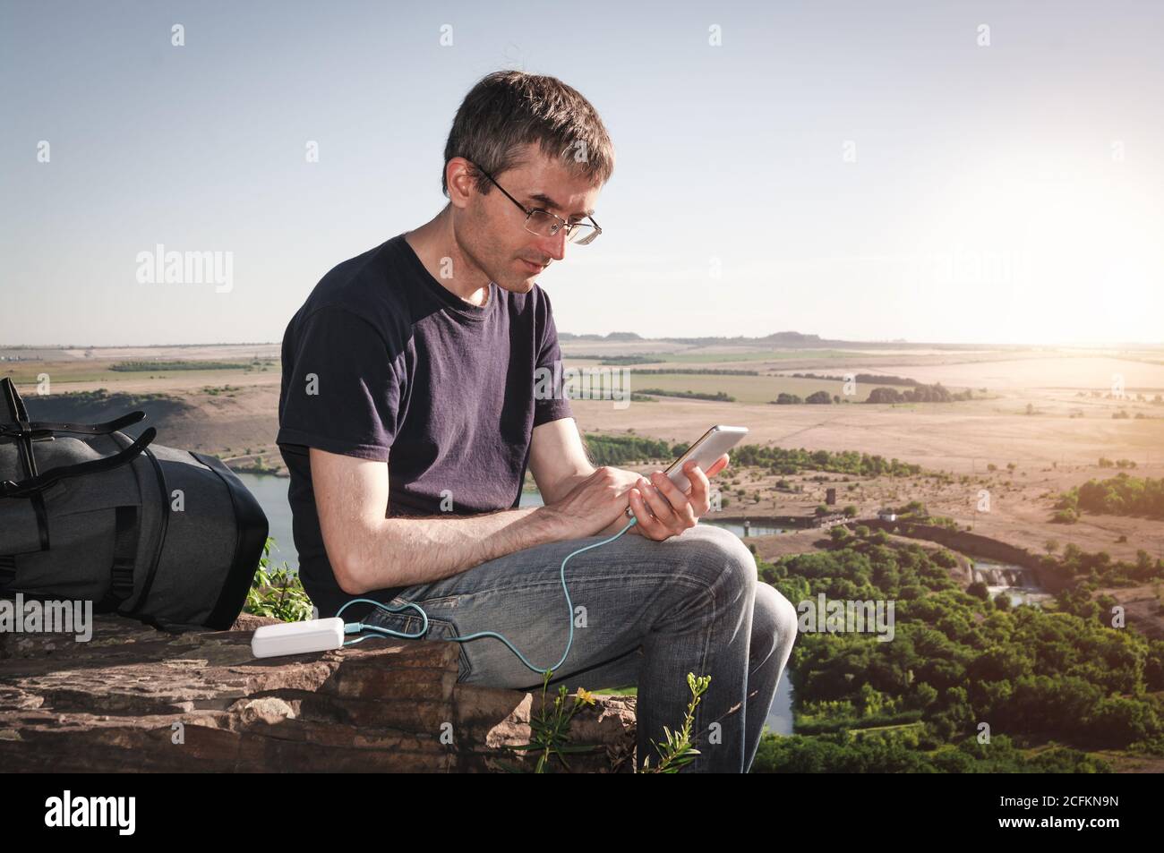 Der Mann auf einer Wanderung nutzt das Smartphone, während er im Morgengrauen von der Powerbank auf dem Felsen lädt. Stockfoto