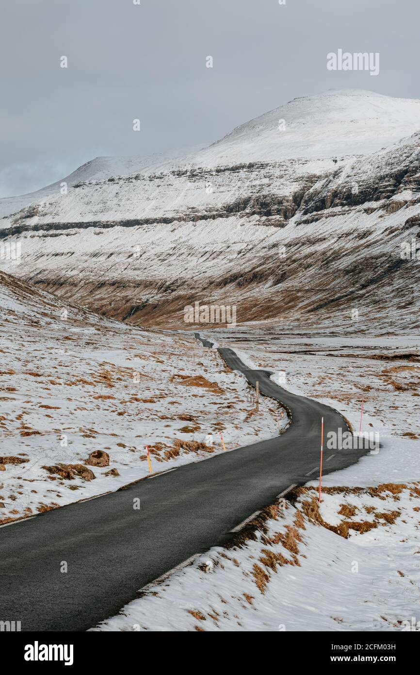 Leere Asphaltstraße mit Schnee bedeckt und von Bergen umgeben An bewölktem Tag auf den Färöern Stockfoto