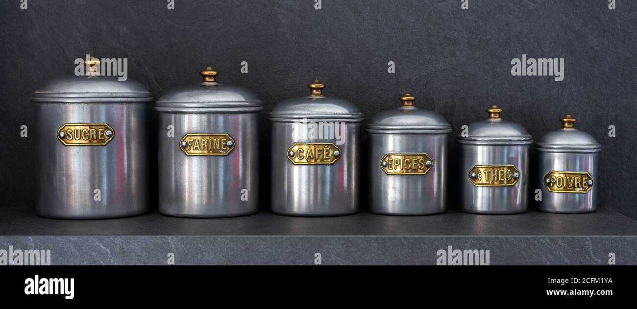 Set aus sechs Zinngläsern in verschiedenen Größen, aufgereiht auf einem Regal mit den Aufschriften 'SUGAR', 'MEHL', 'KAFFEE', 'SPICES', 'TEE' und 'PFEFFER'. Stockfoto