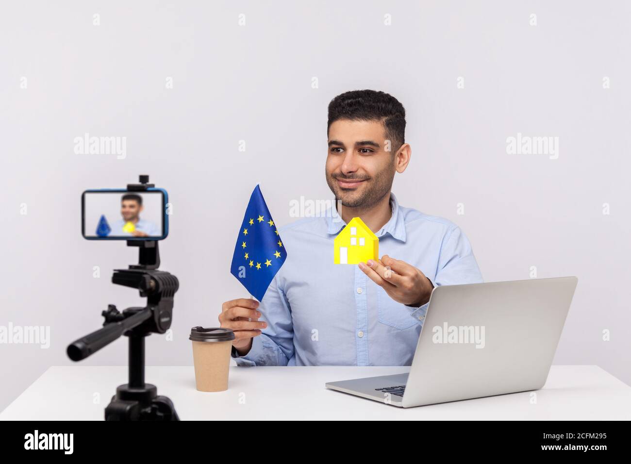 Fröhlicher lächelnder Mann, Immobilienmakler zeigt Papierhaus und EU-Flagge zur Kamera, Video-Aufzeichnung, Streaming Online-Blog über Kauf o Stockfoto
