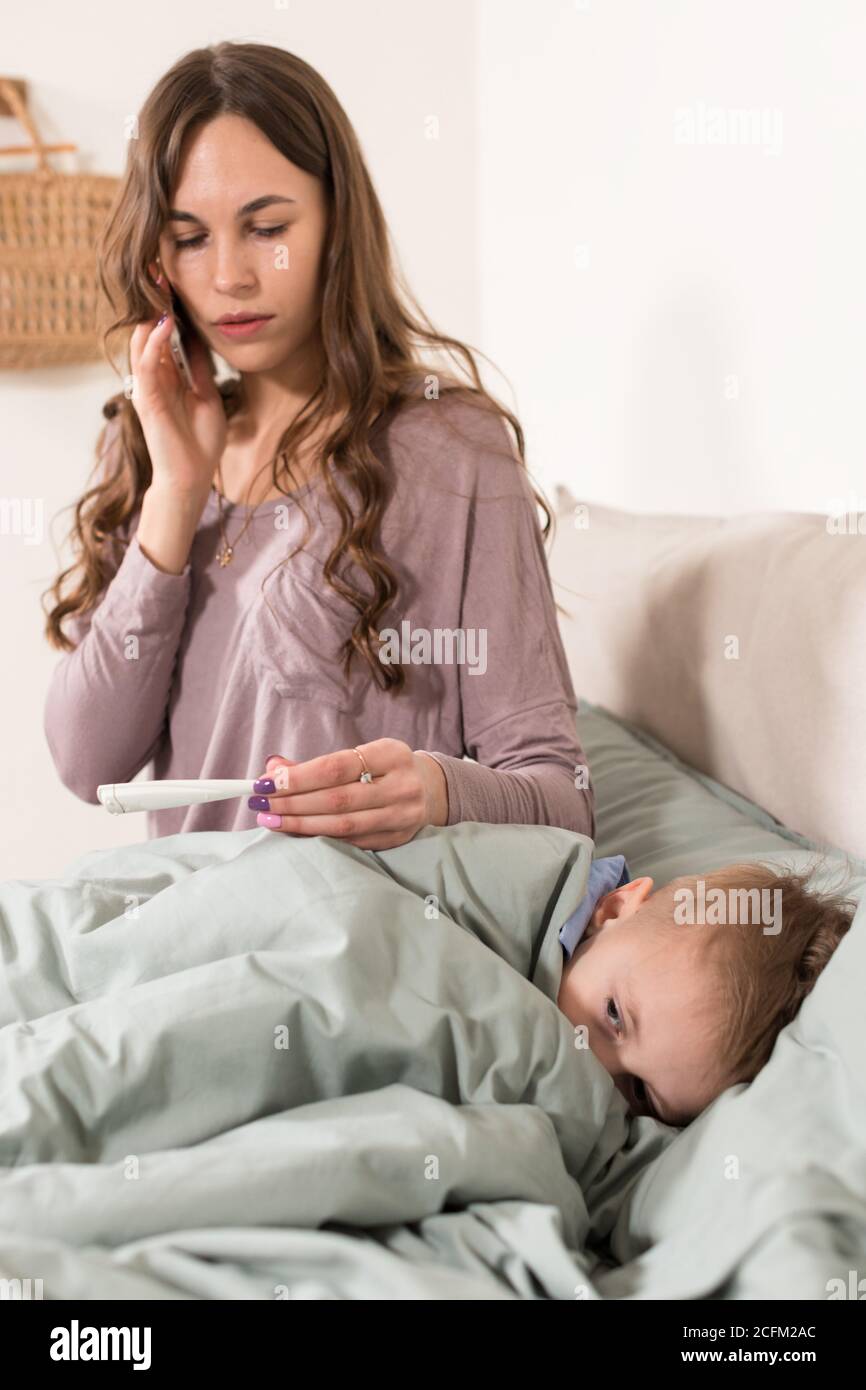 Mutter misst die Temperatur ihres Sohnes und ruft einen Arzt. Krankes Kind fühlt sich schlecht und liegt im Bett unter einer Decke zu Hause. Pandemisches Coronavirus, covid Stockfoto
