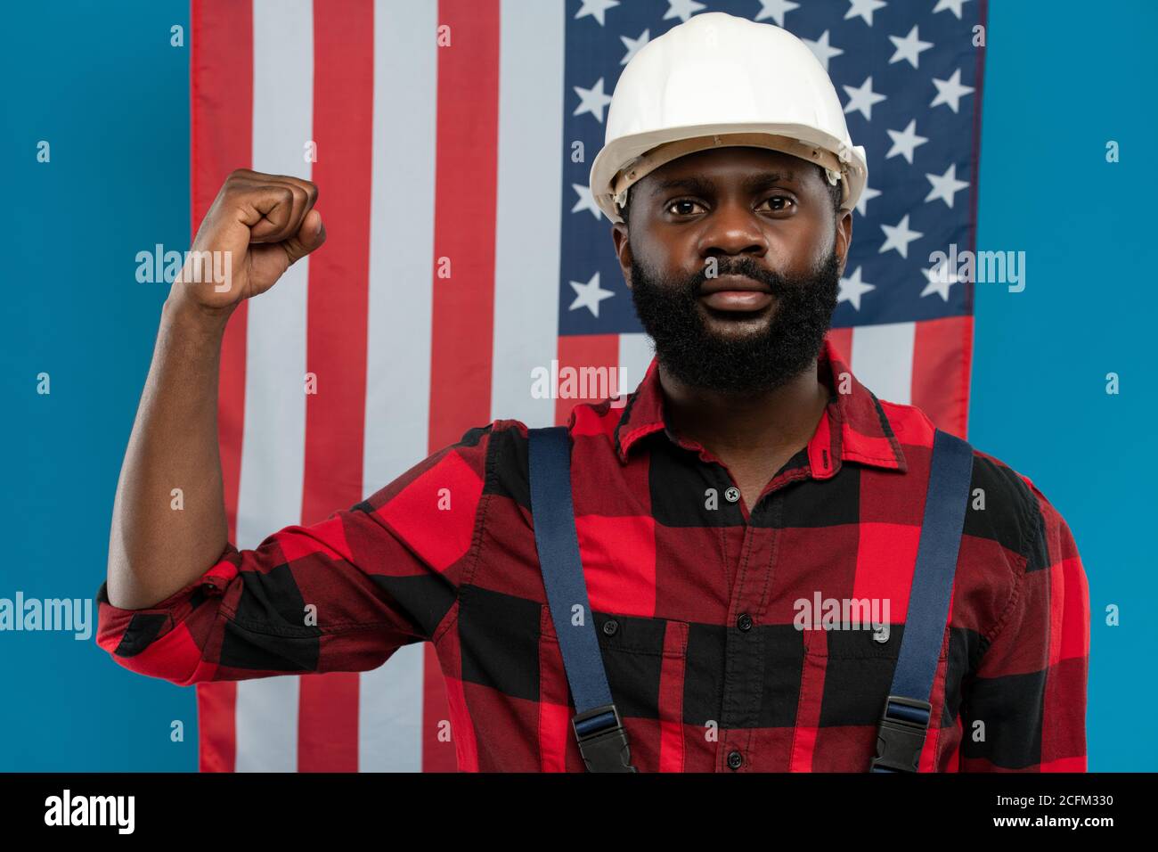 Junger Baumeister oder Vorarbeiter, der seinen starken Arm eingebogen zeigt Ellenbogen gegen US-Flagge Stockfoto