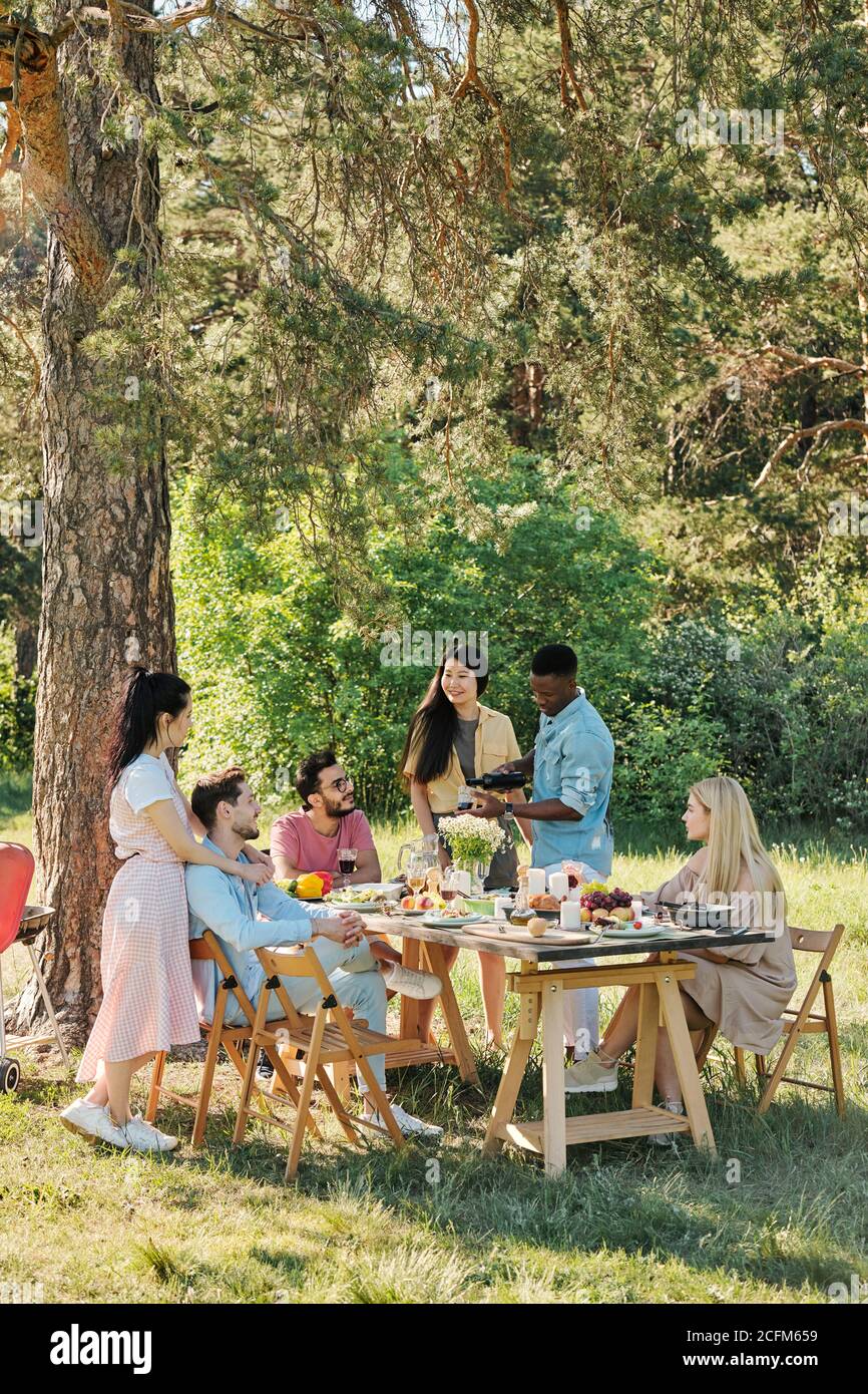 Erholsame junge Freunde sitzen und stehen am Tisch unter serviert Kiefernbaum Stockfoto