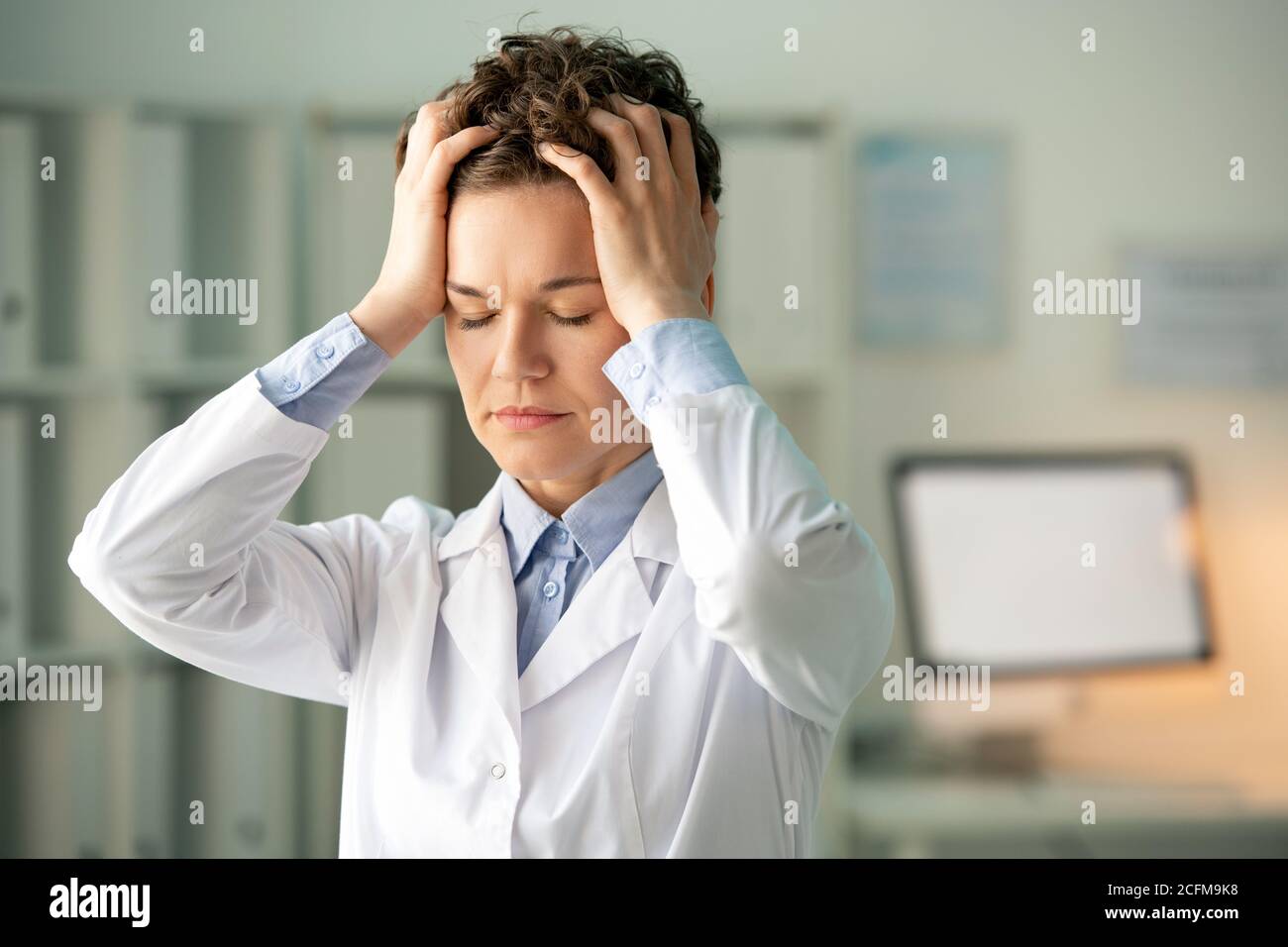 Junge müde Chemikerin oder Apothekerin in Weißkochspüllchen berühren sie Kopf Stockfoto